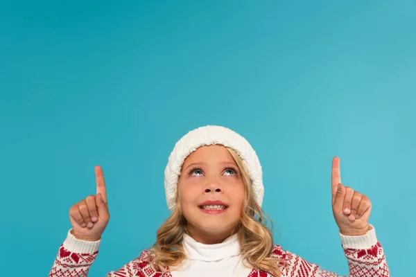 Smiling girl in warm hat looking up and pointing with fingers isolated on blue — Stock Photo