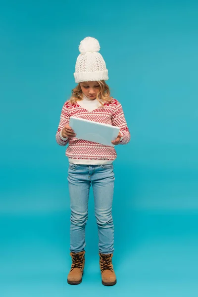Vista a figura intera della ragazza in jeans, cappello a maglia e maglione elegante con tablet digitale su blu — Foto stock