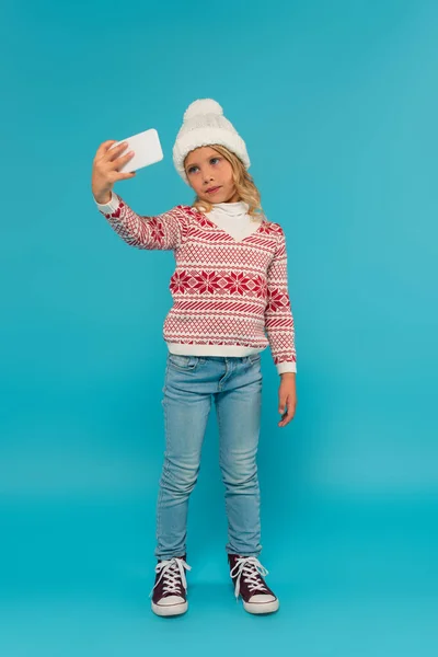Вид в полный рост девушки в теплом свитере и джинсах, делающей селфи на смартфон синего цвета — стоковое фото