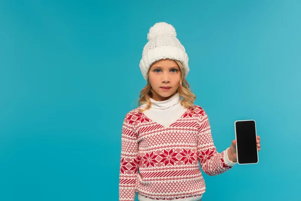 Niño en sombrero de punto y suéter con adorno que muestra el teléfono inteligente con pantalla en blanco aislado en azul - foto de stock