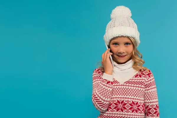 Chica sonriente en sombrero caliente y suéter hablando en el teléfono inteligente aislado en azul - foto de stock