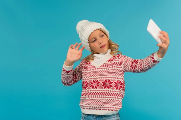 Enfant en bonnet tricoté et pull agitant la main tout en prenant selfie isolé sur bleu — Photo de stock