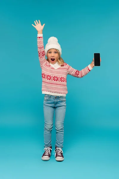 Visão de comprimento total da menina em camisola quente e jeans mostrando celular com tela em branco no azul — Fotografia de Stock