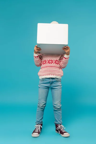 Bambino in maglione caldo e jeans che oscurano il viso con computer portatile su blu — Foto stock