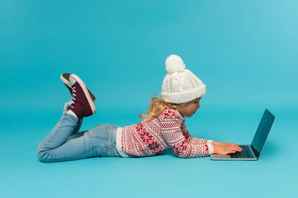 Vista lateral da menina em chapéu, suéter e jeans digitando no laptop com tela em branco no azul — Fotografia de Stock