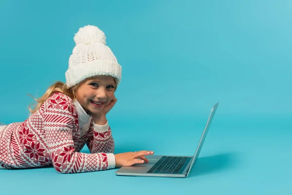 Fille gaie en bonnet tricoté et pull avec ornement à l'aide d'un ordinateur portable tout en étant couché sur bleu — Photo de stock