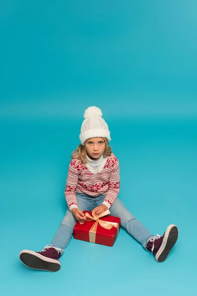 High angle view of girl in knitted hat and sweater looking at camera while sitting near gift box on blue — Stock Photo