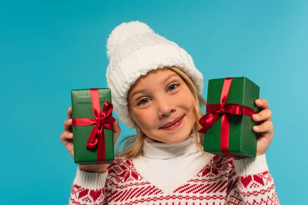 Heureux fille en bonnet tricoté tenant des boîtes-cadeaux vertes isolé sur bleu — Photo de stock