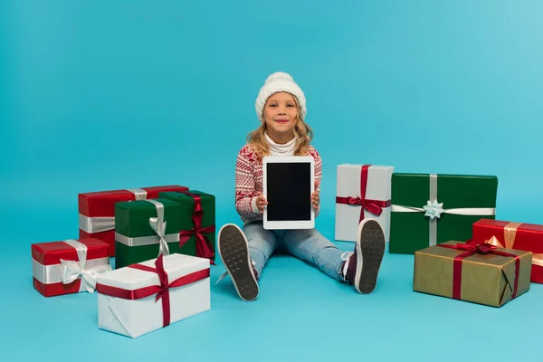 Happy kid in jumper and hat sitting with digital tablet near multicolored gift boxes on blue — Stock Photo
