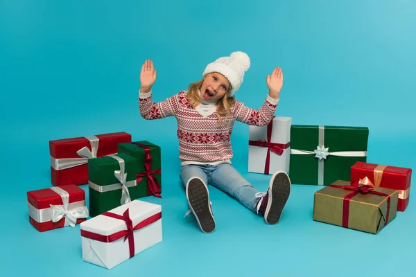 Aufgeregtes Kind gestikuliert und blickt in die Kamera, während es neben bunten Geschenkboxen auf blauem Grund sitzt — Stockfoto
