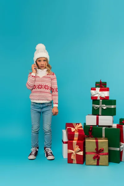 Élégant enfant en chapeau chaud, pull, jeans et gommes parler sur smartphone près de cadeaux sur bleu — Photo de stock