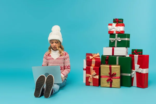 Enfant en bonnet tricoté et pull assis près de la pile de cadeaux et en utilisant un ordinateur portable sur bleu — Photo de stock