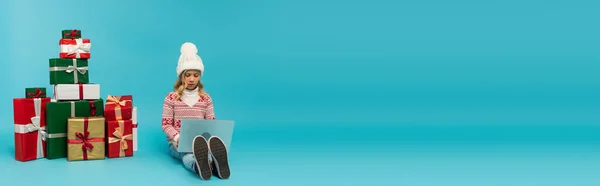 Enfant dans un chapeau chaud à l'aide d'un ordinateur portable tout en étant assis près de boîtes-cadeaux multicolores sur bleu, bannière — Photo de stock