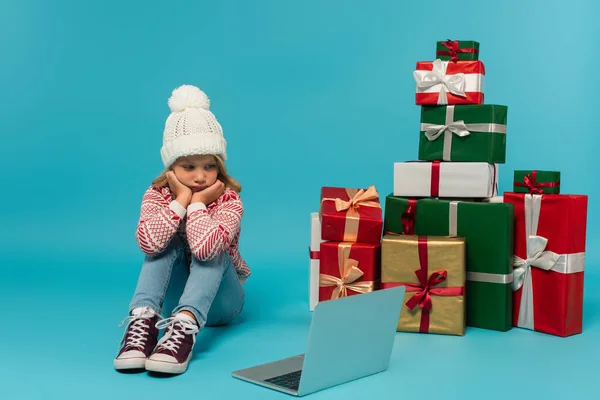 Pensive girl sitting near laptop and stack of multicolored gift boxes on blue — Stock Photo