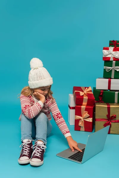 Mädchen in warmem Hut und Pullover mit Laptop, während sie neben bunten Geschenkboxen auf blauem Grund sitzt — Stockfoto