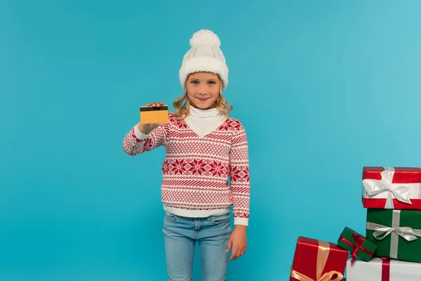 Chica feliz en sombrero de punto y jersey con tarjeta de crédito cerca de cajas de regalo multicolores aislados en azul - foto de stock