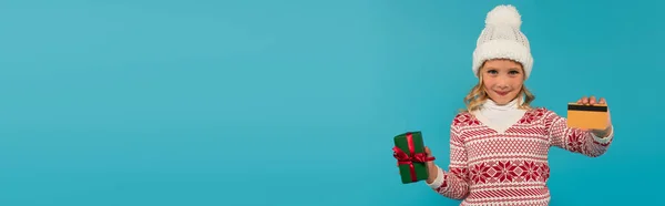 Lächelndes Kind in warmem Hut und Pullover mit Kreditkarte und Geschenk isoliert auf blauem Banner — Stockfoto