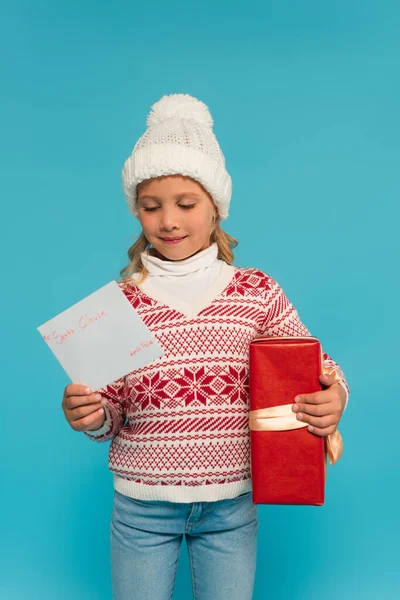 Pleased child in warm sweater and hat holding present and letter to santa clause isolated on blue — Stock Photo