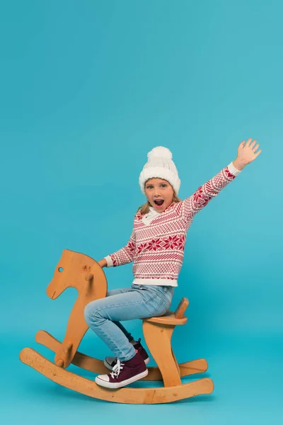 Bambino eccitato in maglia cappello e maglione urlando e agitando mano sul cavallo a dondolo su blu — Foto stock