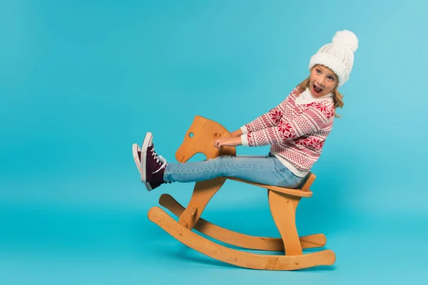 Amazed kid in sweater and hat screaming while riding rocking horse on blue — Stock Photo