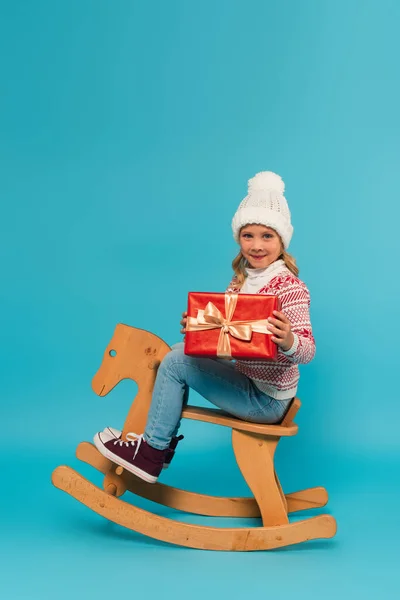Joyful girl in knitted hat and sweater holding present while riding rocking horse on blue — Stock Photo
