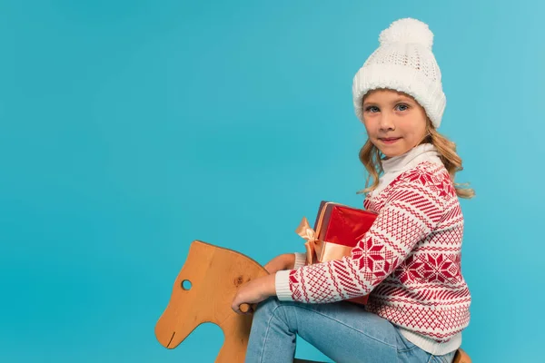 Lächelndes Kind mit warmem Hut reitet Schaukelpferd mit Geschenkbox und lächelt isoliert in die Kamera auf blauem Grund — Stockfoto