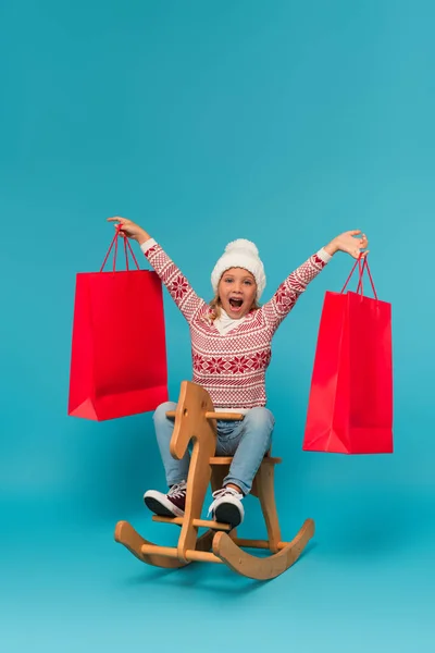 Aufgeregtes Mädchen mit Einkaufstaschen reitet Schaukelpferd auf blau — Stockfoto