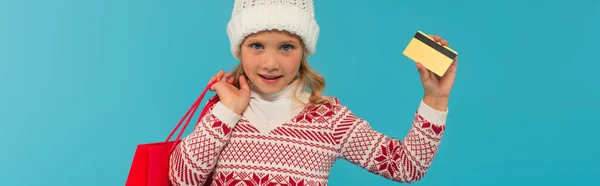 Smiling kid in warm hat holding credit card and shopping bag isolated on blue, banner — Stock Photo