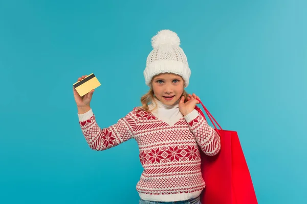 Kid in knitted sweater and hat standing with credit card and shopping bag isolated on blue — Stock Photo