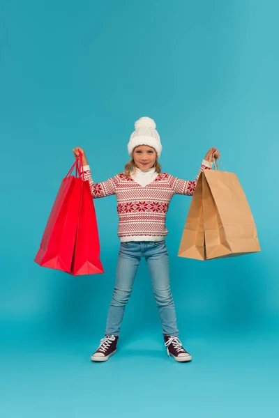 Enfant en pull chaud, jeans et baskets debout avec des sacs à provisions sur bleu — Photo de stock