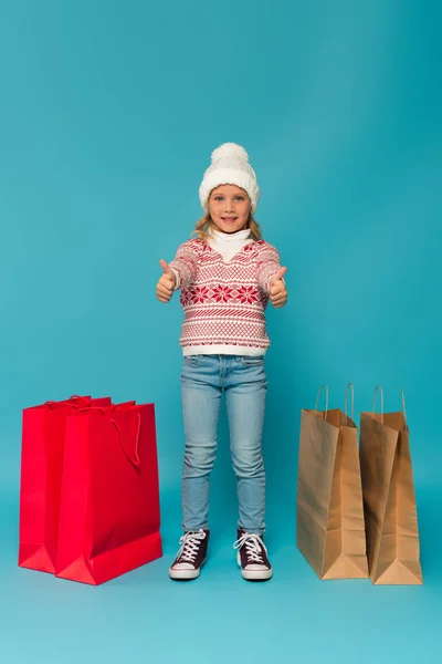 Chica feliz en suéter caliente y pantalones vaqueros mostrando los pulgares hacia arriba cerca de las bolsas de compras en azul - foto de stock