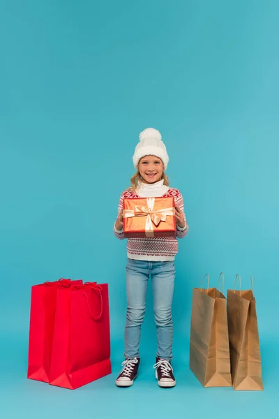 Весёлый ребёнок в трикотажной шляпе и свитере, держащий подарок рядом с пакетами для покупок на голубом — стоковое фото