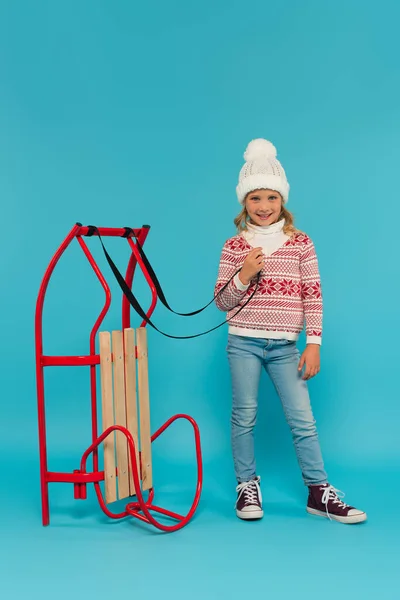 Cheerful girl in warm hat, sweater and jeans standing near sled on blue — Stock Photo