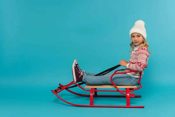 Kid in knitted hat and sweater sitting in sled and looking at camera on blue — Stock Photo