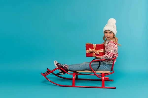 Fille en bonnet tricoté et pull tenant boîte cadeau rouge tout en étant assis en traîneau sur bleu — Photo de stock