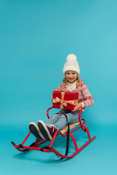 Allegro bambino in slitta che tiene scatola regalo rossa mentre guarda la fotocamera su blu — Foto stock