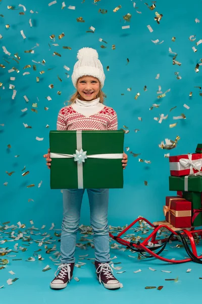 Niño alegre en sombrero caliente sosteniendo gran caja de regalo cerca de confeti y trineo con regalos en azul - foto de stock