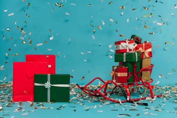 Schlitten mit Geschenken in der Nähe von Einkaufstaschen und Geschenkboxen unter herabfallendem Konfetti isoliert auf blau — Stockfoto