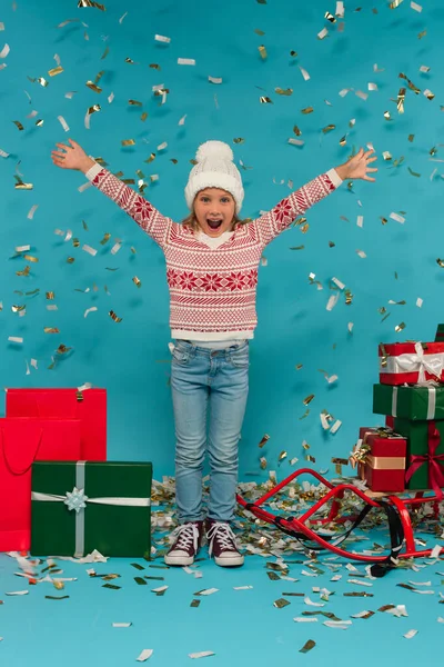 Stylisches und staunendes Kind mit ausgestreckten Händen in der Nähe von Schlitten und Geschenkboxen auf blau — Stockfoto