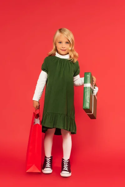 Vista a tutta lunghezza di bambino elegante con scatola regalo e shopping bag su rosso — Foto stock