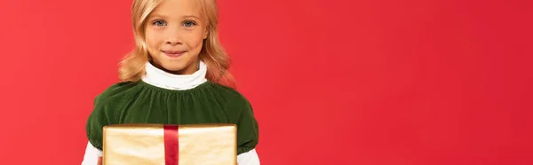 Niño positivo sosteniendo presente mientras sonríe a la cámara aislada en rojo, pancarta - foto de stock