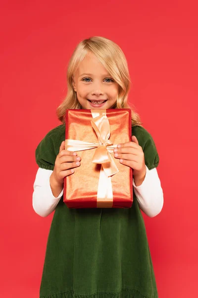 Alegre chica en vestido verde sosteniendo regalo en papel de regalo de oro aislado en rojo - foto de stock