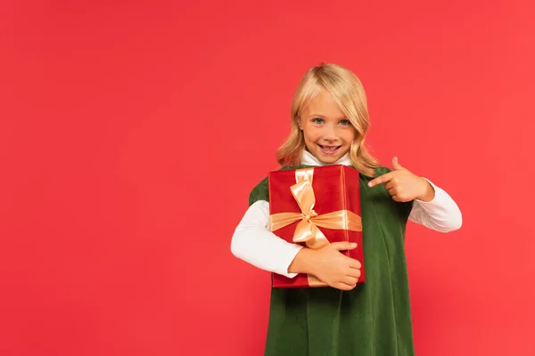 Pleased girl pointing at gift box while smiling at camera isolated on red — Stock Photo