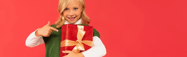 Cheerful kid pointing with finger at gift box isolated on red, banner — Stock Photo