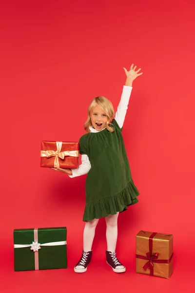 Fille étonnée tenant présent et criant tout en se tenant debout avec la main levée près de boîtes-cadeaux sur rouge — Photo de stock