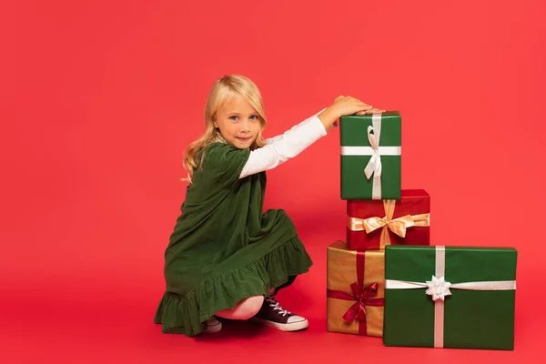 Chica en vestido verde sonriendo a la cámara cerca de pila de cajas de regalo en rojo - foto de stock