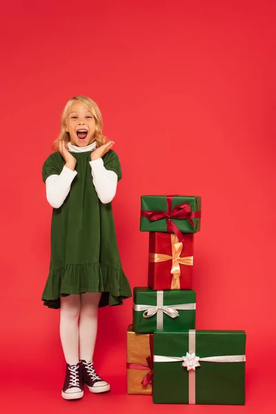 Erstauntes Kind in grünem Kleid und Gummischuhen klatscht Hände neben Stapel von Geschenken auf rot — Stockfoto