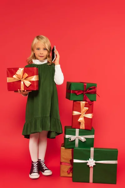 Visão de comprimento total da menina em vestido verde chamando no smartphone perto de caixas de presente no vermelho — Fotografia de Stock