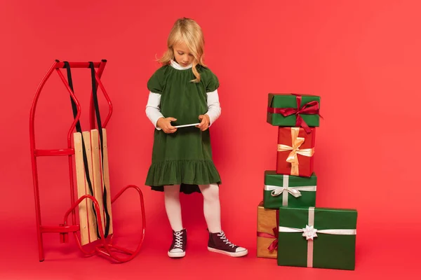 Fille en robe élégante en utilisant un téléphone mobile près traîneau et pile de boîtes-cadeaux sur rouge — Photo de stock