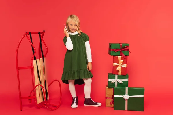 Улыбающаяся девушка в зеленом платье и ботинки, говорящие на смартфоне рядом с подарками и сани на красном — стоковое фото